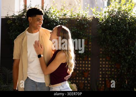 Diverse couple standing in sunlit backyard garden by wooden trellis, smiling, smartwatch visible Stock Photo