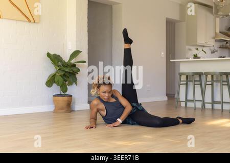 Adult African woman performing side-floor leg raise on light wood floor, wearing athletic top Stock Photo