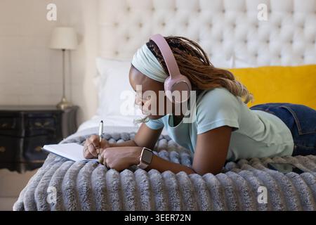 African American woman lying on stomach on bed writing in notebook with pen and pink headphones Stock Photo