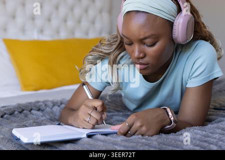 Adult African woman in bedroom lying on bed writing in notebook wearing pink headphones, smartwatch Stock Photo