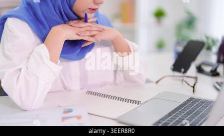 Muslim businesswoman in blue hijab thinking while working at modern office desk with laptop and notebook documents Stock Photo