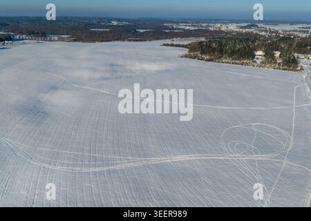 Aerial view of a vast snow-covered field with intricate tracks and a distant forest under a clear winter sky in Lithuania. Stock Photo