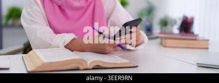 Muslim woman in hijab using smartphone while studying with books and taking notes at desk for education and learning Stock Photo