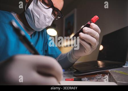 Healthcare worker wearing protective mask and gloves examining a blood vial and writing notes Stock Photo
