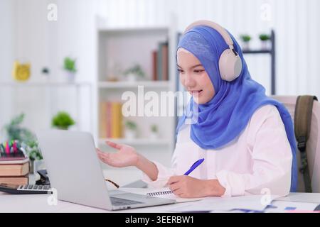 Young Muslim Asian women wearing blue hijab and headphones having online learning session using laptop computer at home Stock Photo