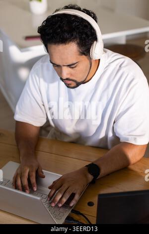 Mid-adult African American man typing on laptop at home table, wearing headphones and smartwatch Stock Photo
