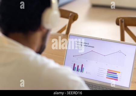 African American man in mid 30s analyzing dashboard on laptop at wooden table wearing headphones Stock Photo