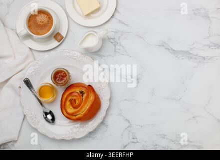 A sweet bun with raisins, jam and honey in a white plate on a marble background for breakfast. Coffee, milk, butter on the table. Selective focus, top Stock Photo
