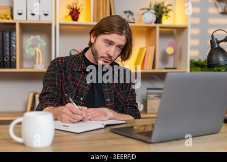 Young man on home table watches study video on laptop stops playback and writes ideas in notebook. Adult guy on home table stays focused on online learning and keeps taking notes with calm attention. Stock Photo