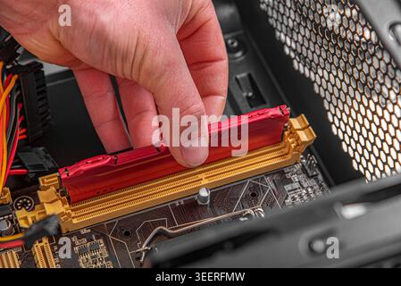 A technician's hand is shown installing a red RAM module into a motherboard slot inside a computer case. Stock Photo