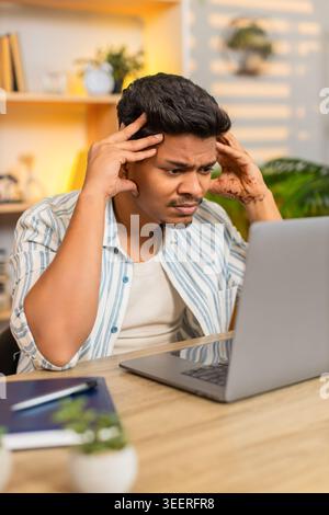Indian man on home table looks at laptop screen with focused mind while solving work task in living room. Arabian guy on home table thinks hard about work project and plans next steps with calm focus. Stock Photo