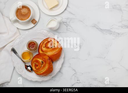Two buns with raisins, jam and honey in a white plate on a marble background for breakfast. Coffee, milk, butter on the table. Selective focus, top vi Stock Photo