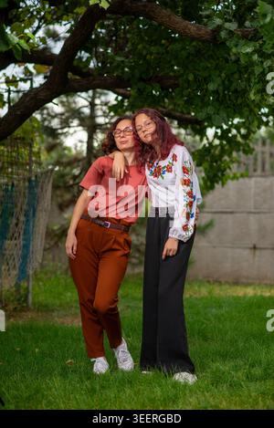 Two women wearing glasses pose together outdoors under a tree, showing friendship and casual fashion. Stock Photo