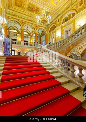 People move up the red carpet stairs in the Hungarian State Opera. The hall has bright lights and ornate designs on the walls. Stock Photo