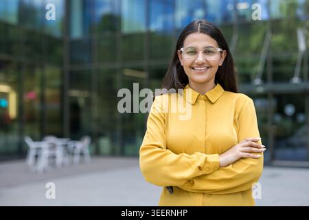 Young professional woman crossing arms, wearing clear frame glasses and a yellow shirt, smiling outside a modern glass office building, portraying confidence and success Stock Photo