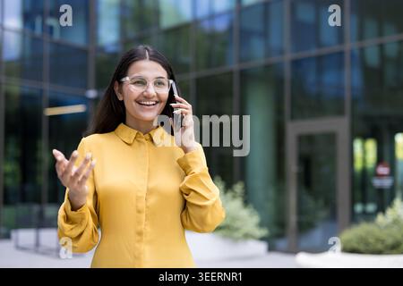 Indian businesswoman smiling confidently while having a professional conversation on her mobile phone outside a modern office building, representing corporate communication and success Stock Photo