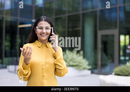 Smiling businesswoman having a phone conversation outdoors, communicating with clients or colleagues while standing confidently in front of a contemporary glass office building Stock Photo