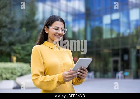 Smiling young businesswoman wearing clear glasses and a yellow shirt, standing outside a modern office building, holding and looking at a digital tablet Stock Photo