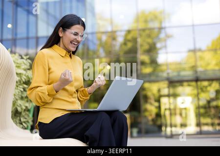 Excited young professional woman in glasses sitting outdoors, joyfully cheering with clenched fists while watching her laptop screen, feeling triumph and achievement Stock Photo