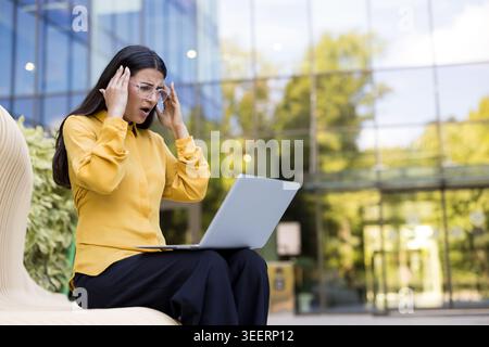 Stressed businesswoman feeling overwhelming headache and frustration, touching her temples while looking at a laptop with a shocked expression, working in front of a modern office building Stock Photo