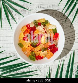 Colorful assorted gummy candies in white bowl surrounded by tropical palm leaves in bright sunlight. Stock Photo