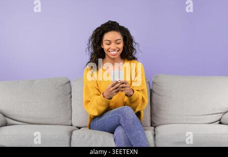 Woman Sitting on Couch Using Smartphone and Smiling in a Bright Room Stock Photo