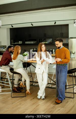 A group of four people engage in a discussion in a contemporary office. Two individuals stand while sharing documents. Two others sit nearby focused o Stock Photo