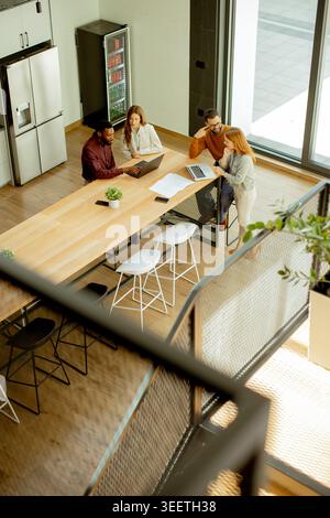 Four people sit at a long table in a modern office. They work on laptops and discuss ideas. Sunlight comes through windows, casting light on the room. Stock Photo