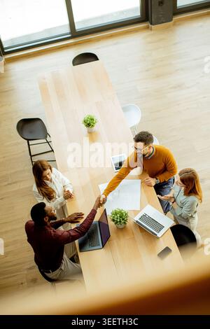 A group of four people meet in a modern office setting. Two individuals shake hands at a long wooden table. Laptops and documents are present as they Stock Photo