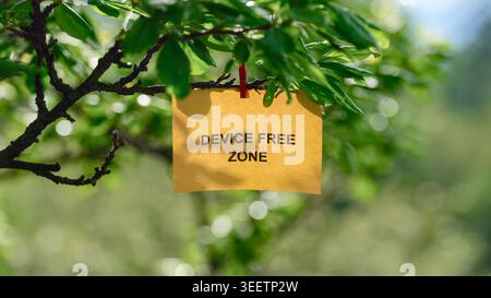 A yellow note with the text Device Free Zone on it clipped to a green tree branch, representing a digital detox and mindfulness concept. Close up. Stock Photo
