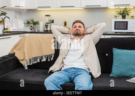 Young man at home leans back and places hands behind head and relaxes muscles with slow breath. Caucasian guy on sofa stretches elbows widens chest and shows calm relief smile and steady mood again Stock Photo
