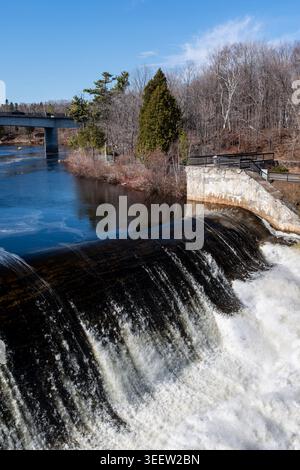 Visitors to Montmorency Falls park look at the upper level of the waterfall. Warm early spring sun and forest frame the scene beneath blue sky. Stock Photo