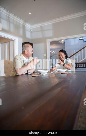 Asian couple sitting at dining table in home, holding glasses, laughing near chandelier Stock Photo