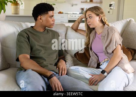 Couple sitting on three-seat sofa in living room, talking while showing smartwatch and bracelets Stock Photo
