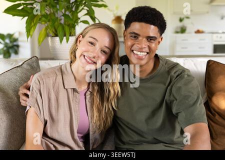 Diverse couple sitting on light sofa in bright living room with potted plant, wearing casual wear Stock Photo