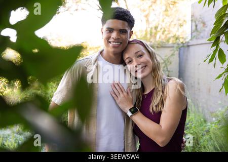 Diverse couple standing in sunlit backyard, smiling, woman wearing smartwatch and maroon top Stock Photo