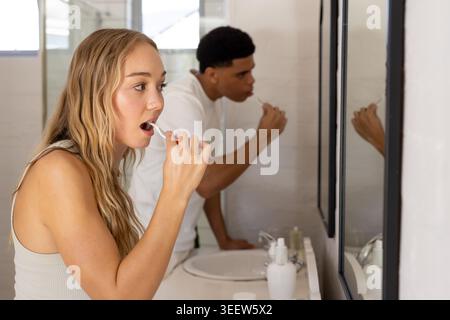 Couple brushing teeth at bathroom sink, blonde woman holding toothbrush, man leaning by mirror Stock Photo