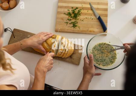 Couple slicing loaf with serrated knife and whisking herb eggs in glass bowl on kitchen countertop Stock Photo