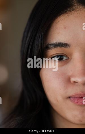 Asian female posing in softly lit interior showing tight half-face with light reflection, dark top Stock Photo