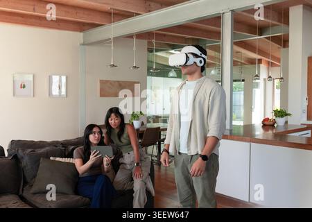 Diverse friends watching man wearing white VR headset near kitchen, women holding tablet on sofa Stock Photo