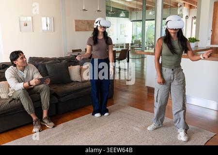 Diverse friends wearing white VR headsets and holding controllers in modern living room with tablet Stock Photo