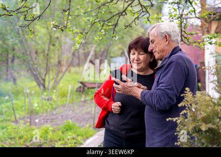 Senior Couple Sharing Smartphone in Spring Garden. Older couple looking at a smartphone together in a blooming garden, enjoying technology, connection Stock Photo