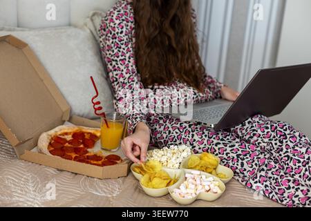 Woman Relaxing with Laptop and Snacks. Young woman in pajamas using a laptop on bed with pizza, juice, popcorn, chips, and sweets for cozy Stock Photo