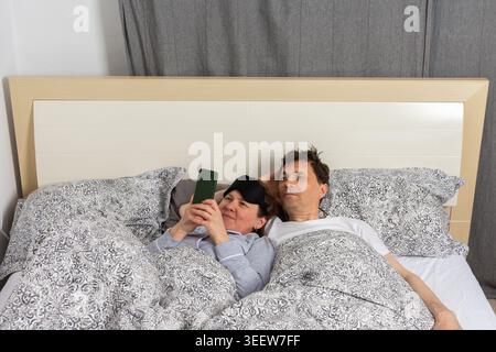 A married couple wakes up in bed on a typical morning. The woman is using a phone while the man lies next to her. They share a cozy moment in their be Stock Photo