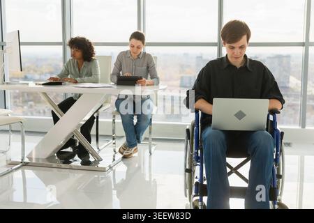 Accessible environment. Young man in chair for people with disability working with laptop in modern office. Diverse multiracial group of people colleagues working together. Young professional team Stock Photo