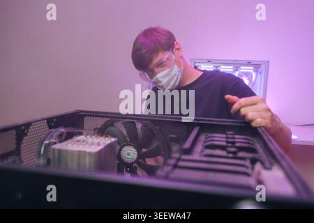 Technician in face mask and safety glasses assembles desktop PC. Low angle view from inside computer case during hardware repair or upgrade. Purple ne Stock Photo