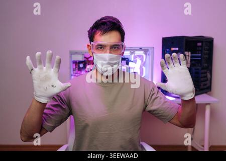 Portrait of young male technician in protective mask and glasses showing dirty gloves. He just finished assembling or cleaning computer. Modern Stock Photo
