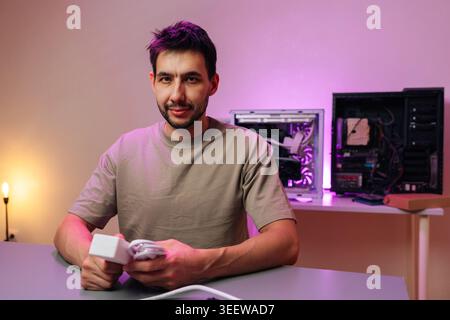 Confident technician holding power adapter at desk. Open PC cases in background with purple neon light. IT support, hardware repair, custom computer Stock Photo