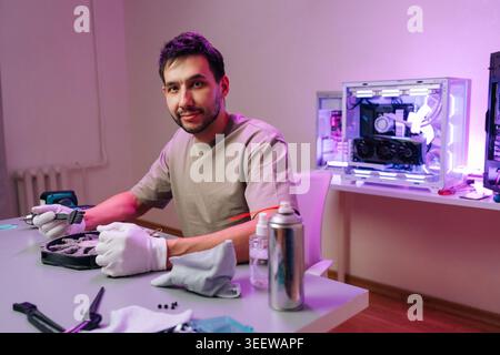 Smiling technician cleaning dusty computer fan with brush. Man in gloves at desk performs PC maintenance. Workshop has custom computer with purple Stock Photo