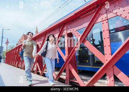 Young diverse couple holding hands and walking on red metal bridge in city near blue tram Stock Photo